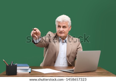 Portrait of senior teacher with laptop at table against green chalkboard