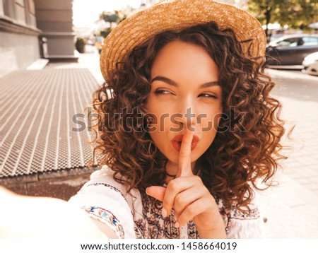 Beautiful model with afro curls hairstyle dressed in summer hipster dress and hat.Carefree girl posing in the street.Taking selfie self portrait photos on smartphone.Finger showing hush silence sign