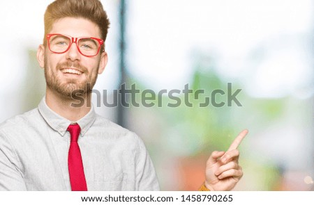 Young handsome business man wearing glasses with a big smile on face, pointing with hand and finger to the side looking at the camera.