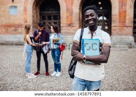 Young cheerful successful african nerdy student standing with books, his classmates on the background, park near campus, sunny day .