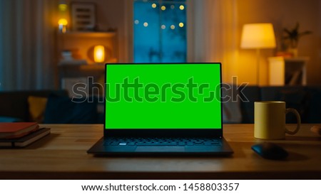 Laptop Computer Showing Green Key Screen Stands on a Desk in the Living Room. In the Background Cozy Living Room in the Evening with Warm Lights on.