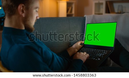 Man Sitting Relaxes on a Couch Works on a Laptop with Green Key Screen. Late at Night in His Living Room Man Uses Notebook Computer.