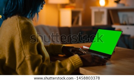 Beautiful Black Woman Sitting at Her Desk Works on a Laptop with Green Mock-up Screen. Late at Night in Her Living Room Girl Uses Notebook Computer.