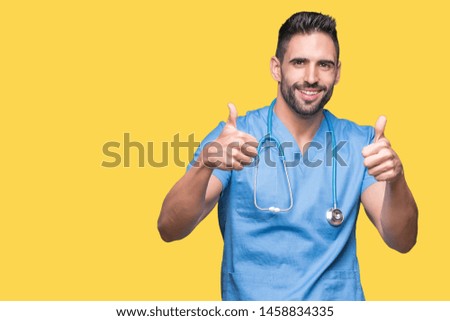 Handsome young doctor surgeon man over isolated background success sign doing positive gesture with hand, thumbs up smiling and happy. Looking at the camera with cheerful expression, winner gesture.