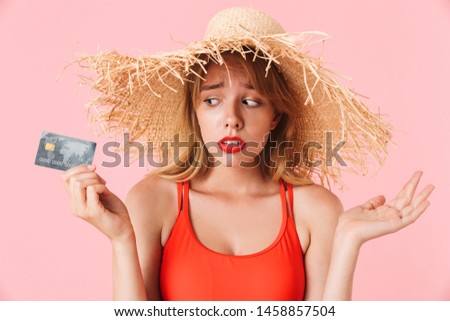 Portrait of confused young woman with long curly hair wearing summer straw hat holding credit card isolated over pink background