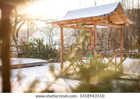 Beautiful wooden small fairy elf arbor gazebo in the sunlight dawn's sunshine in winter snow against the background of spruce pine forest