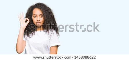 Young beautiful girl with curly hair wearing casual white t-shirt smiling positive doing ok sign with hand and fingers. Successful expression.