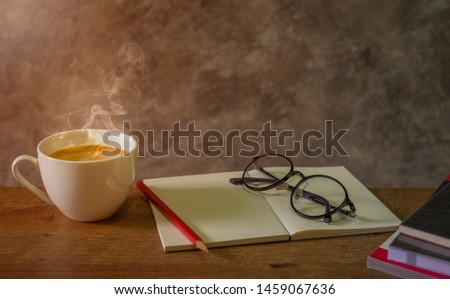 Hot white cup of fresh coffee, cappuccino on the wooden table with Blank open booklet, red pencil, glasses and stack of books to read. morning light from window, Concept education.