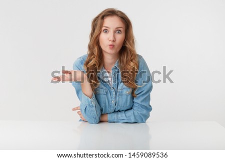 Photo of young cute blonde woman wears in denim shirts, sitting at the white table and sends kiss, looks happy and cheerful, isolated over white background.