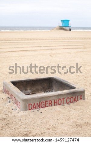 Concrete fire pit with "Danger Hot Coals" sign on a sandy beach, with a life guard tower along the shoreline in the background, and space for text on top