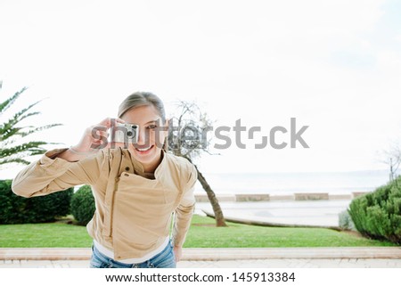 Fashionable attractive young woman using a digital photo camera to point and take pictures near a beach with a green park and the sky in the background.