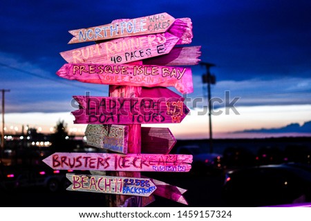 Close up nighttime view of pole with several wooden arrow signs with words naming local Erie Pennsylvania popular locations and the distance to them dark parking lot and sunset background