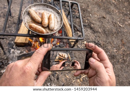 Man takes pictures on phone grilled sausages on pan grill with smoke and flame on a meadow. Barbecue picnic concept