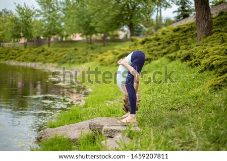 Slim young brunette girl yogi does difficult yoga exercises on the green grass on the background of water.