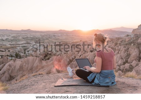 Back view of young woman with laptop sitting on the edge of canyon valley. Remote working concept. 