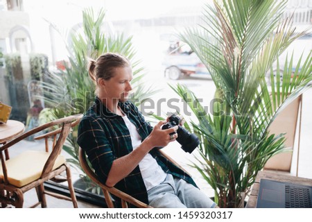 Creative photographer holding professional camera, sitting at cafe with laptop, editing photos.