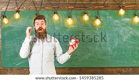 Forgot about time concept. Man with beard and mustache on shocked face in classroom. Teacher in eyeglasses holds alarm clock. Bearded hipster holds clock, chalkboard on background, copy space.