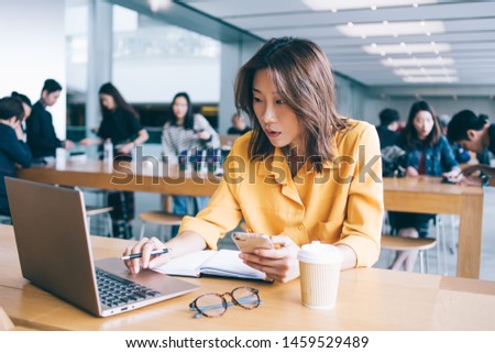 Concentrated Asian female freelancer in yellow shirt working typing on laptop and using smartphone at desk in e-store in Hong Kong city Royalty-Free Stock Photo #1459529489