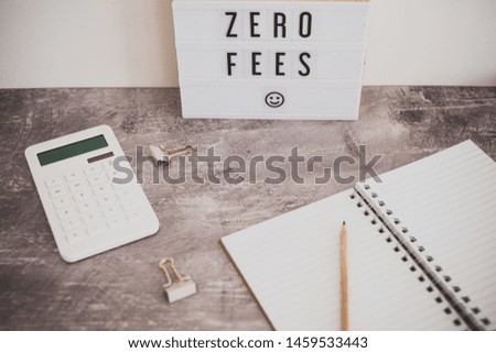 zero fees message on lightbox surrounded by coins calculator on office stationery on concrete desk, muted tones with added vignette effect