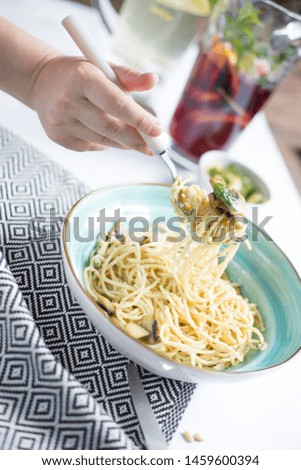 fork with pasta in a female hand near a plate with pasta and mushrooms on a light background of a table.