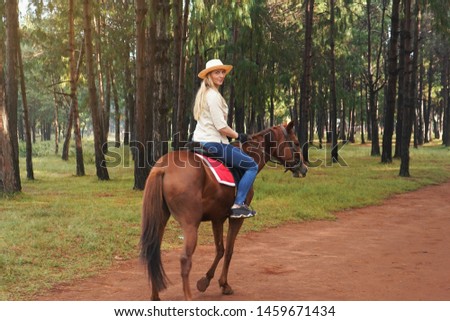 Young woman in shirt and straw hat, riding brown horse in the park looking back over her shoulder, blurred background with trees