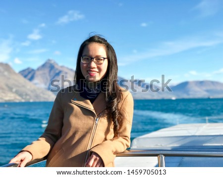 Beautiful Vietnamese girl long curly hair big smile stand on cruise ship light brown winter coat black trouser boots model luxury elegant cruising ship Queenstown New Zealand sunshine happy vacation