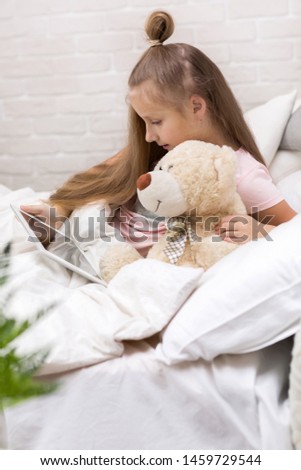 cute little child girl with teddy bear lies in bed uses digital tablet. child playing on tablet pc.