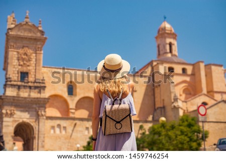 tourist girl with her back to the camera looks and takes a photo of an old building, on a model blue stylish dress and fashionable hat, a small backpack, long blond hair, outdoor, horizontal banner

