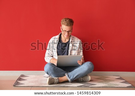 Handsome young man with laptop sitting near color wall