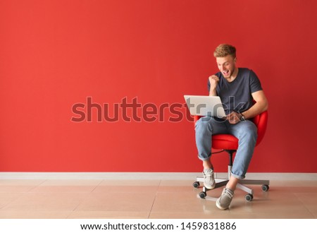 Happy young man with laptop sitting on chair near color wall