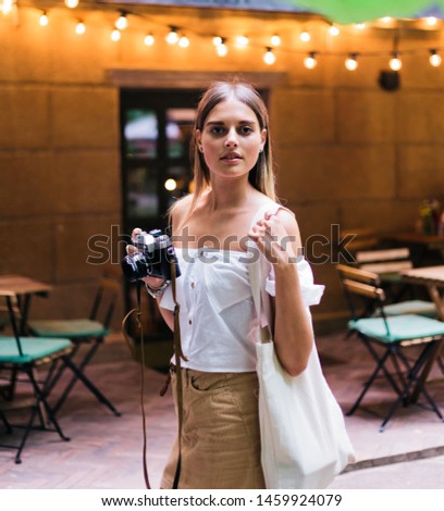 Cheerful blond woman in fashionable clothes holding retro camera in evening on the background of garlands outdoor