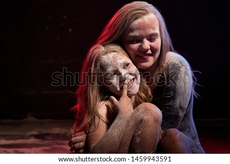 Two sisters in black studo with red and blue light during photoshoot with flour. Small girl and female tennager with white dust on the the face sitting on the floor