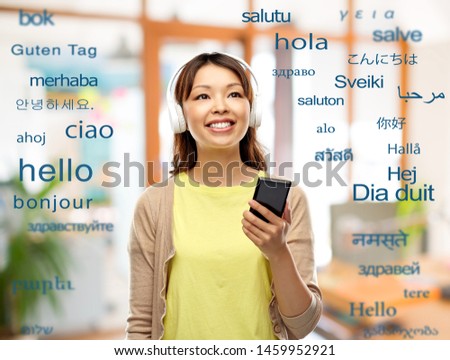 education, communication and technology concept - happy asian young woman in headphones listening to music on smartphone over greeting words in different foreign languages