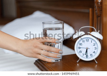 A white alarm clock and a glass of water are on a wooden bedside table. A woman stretches her hand to a glass of water