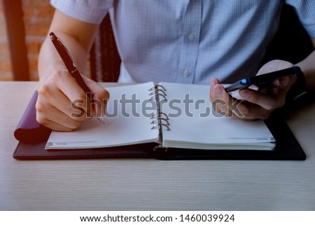 stock photo man writing on blank page note book and holding phone in another hand, notebook placed on wooden table beside window, work at home or coffee shop, copy space, selective focused on hand.