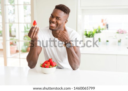 African american man eating fresh healthy strawberries happy with big smile doing ok sign, thumb up with fingers, excellent sign