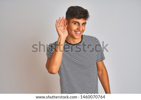 Young indian man wearing navy striped t-shirt standing over isolated white background smiling with hand over ear listening an hearing to rumor or gossip. Deafness concept.