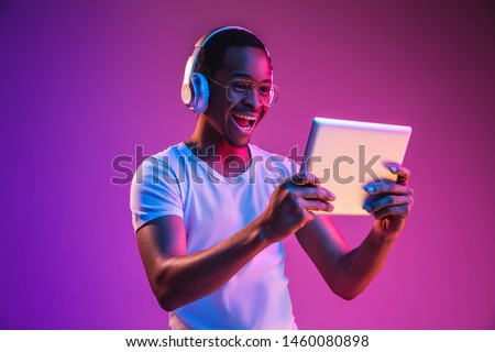 Young african-american man's listening to music in headphones, using tablet in neon light on gradient background. Concept of human emotions, facial expression, summer holidays or weekend, hobby,