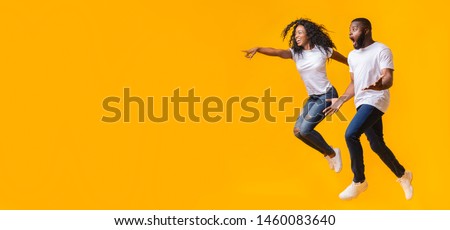 Surprised Black Man Is Looking At Free Space, afro woman is jumping and pointing at empty space, yellow background, panorama Royalty-Free Stock Photo #1460083640