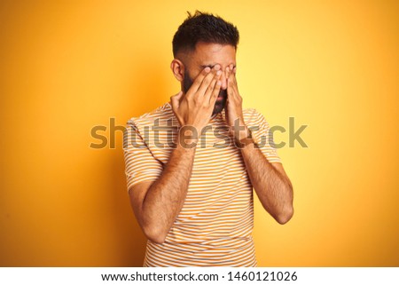 Young indian man wearing t-shirt standing over isolated yellow background rubbing eyes for fatigue and headache, sleepy and tired expression. Vision problem