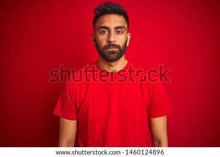 Young handsome indian man wearing t-shirt over isolated red background with serious expression on face. Simple and natural looking at the camera.