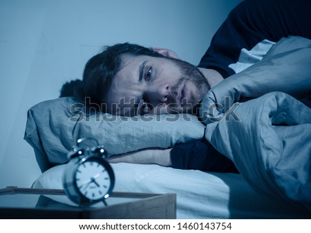Sleepless and desperate young caucasian man awake at night not able to sleep, feeling frustrated and worried looking at clock suffering from insomnia in stress and sleeping disorder concept. Royalty-Free Stock Photo #1460143754