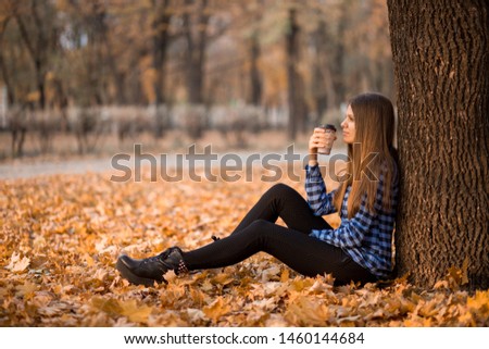 Fall concept. Happy and cheerful woman drinking coffee while sitting on park leaves under fall foliage. Beautiful young modern woman smiling