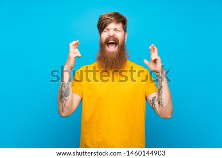 Redhead man with long beard over isolated blue background with fingers crossing