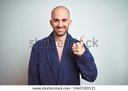 Young man wearing blue bathrobe, relaxed lifestyle over isolated background doing happy thumbs up gesture with hand. Approving expression looking at the camera with showing success.