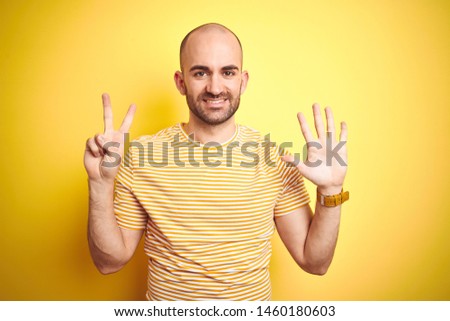 Young bald man with beard wearing casual striped t-shirt over yellow isolated background showing and pointing up with fingers number seven while smiling confident and happy.