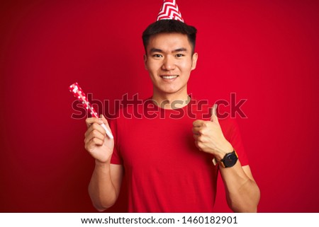 Asian chinese man on birthday celebration wearing funny hat over isolated red background happy with big smile doing ok sign, thumb up with fingers, excellent sign