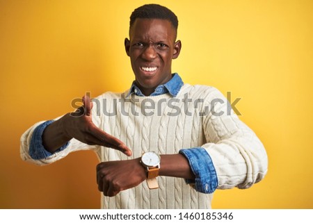 African american man wearing denim shirt and white sweater over isolated yellow background In hurry pointing to watch time, impatience, upset and angry for deadline delay