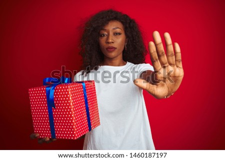 Young african american woman holding birthday gift standing over isolated red background with open hand doing stop sign with serious and confident expression, defense gesture