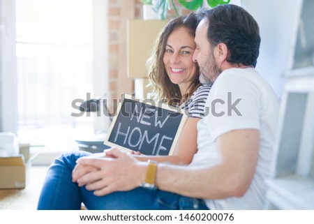 Middle age senior romantic couple in love sitting on the apartment floor with boxes around and showing blackboard smiling happy for moving to a new home